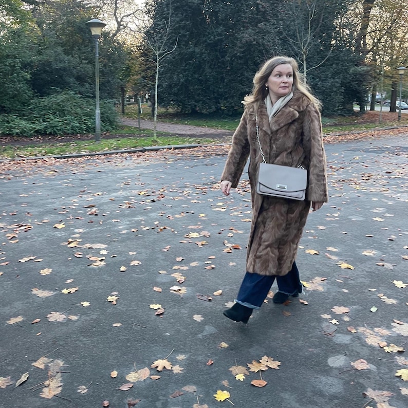 Pu&ograve; includere: Una donna indossa un lungo cappotto di pelliccia marrone, una borsa grigio chiaro, jeans blu scuro e stivali neri. Cammina su un sentiero coperto di foglie. Il cappotto ha un classico colletto a revers e una chiusura con bottoni.