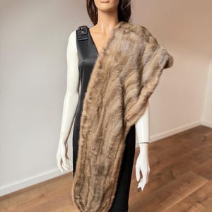 Pode incluir: Uma estola de pele castanha drapeada sobre um vestido preto num manequim. A estola tem um aspeto macio e texturizado com v&aacute;rias tonalidades de castanho. O vestido &eacute; sem mangas com decote em V e um adorno decorativo no ombro.