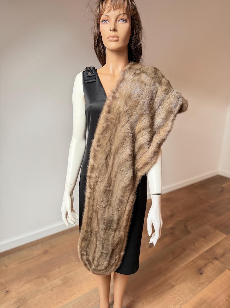 Pode incluir: Uma estola de pele castanha drapeada sobre um vestido preto num manequim. A estola tem um aspeto macio e texturizado com v&aacute;rias tonalidades de castanho. O vestido &eacute; sem mangas com decote em V e um adorno decorativo no ombro.
