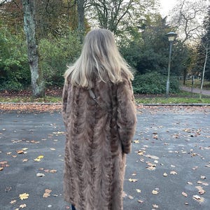 Pu&ograve; includere: Un lungo cappotto di pelliccia marrone con un motivo strutturato. Il cappotto &egrave; indossato da una persona in piedi su una superficie pavimentata con foglie cadute. Il cappotto sembra essere lungo fino al ginocchio e ha uno stile classico ed elegante.