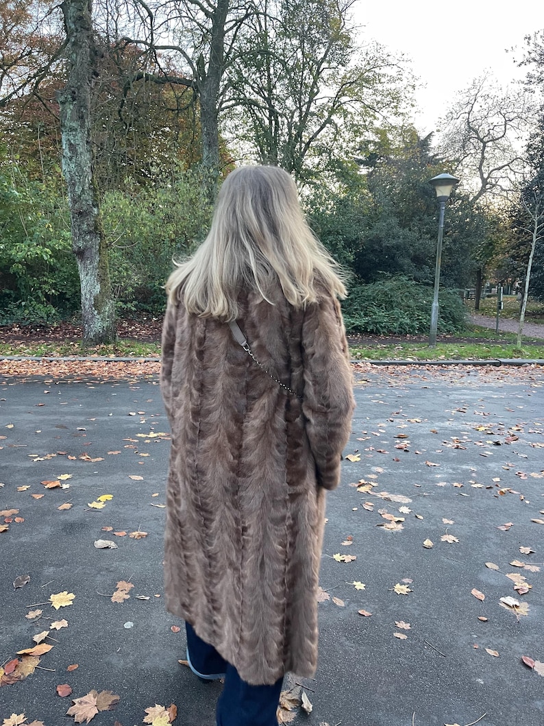 Pu&ograve; includere: Un lungo cappotto di pelliccia marrone con un motivo strutturato. Il cappotto &egrave; indossato da una persona in piedi su una superficie pavimentata con foglie cadute. Il cappotto sembra essere lungo fino al ginocchio e ha uno stile classico ed elegante.