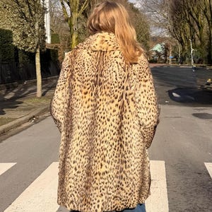 Puede incluir: Un abrigo vintage con estampado de leopardo y cuello grande. El abrigo presenta un patrón de manchas marrones sobre un fondo beige. La persona lleva vaqueros azules. La imagen está tomada al aire libre en un día soleado.