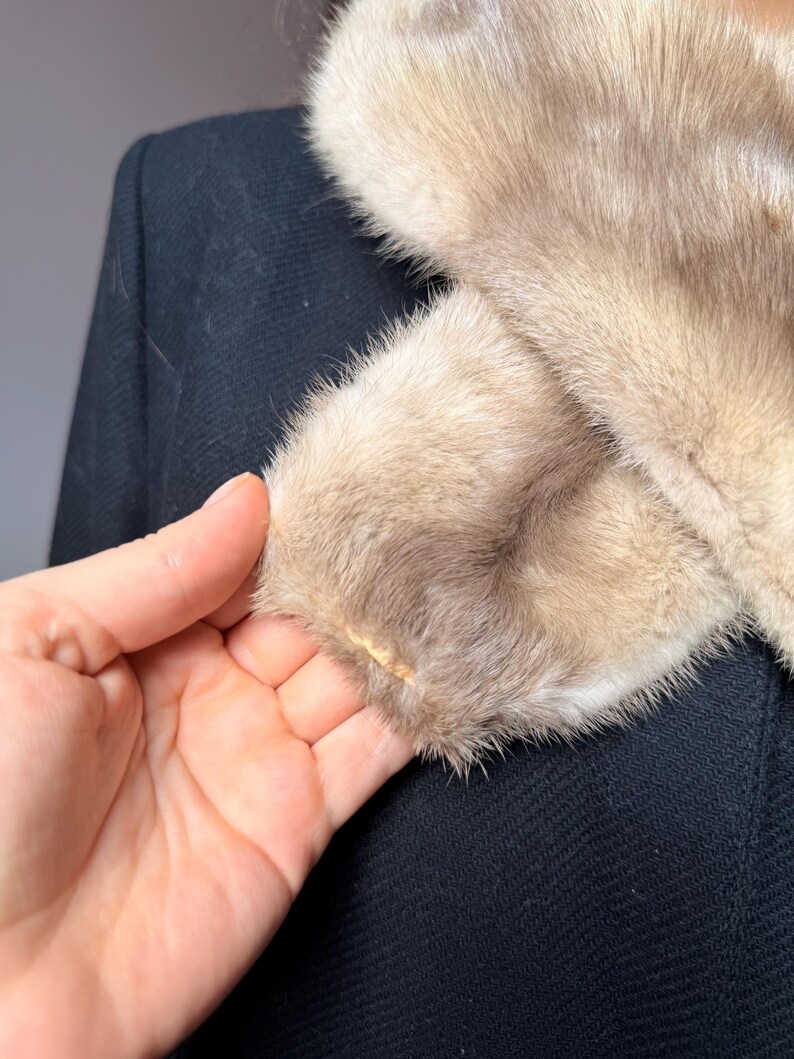 May include: Close-up of a vintage black coat with a light brown fur collar and cuffs. The fur has a soft, textured appearance. The coat's fabric is a dark, woven material.