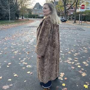 Pu&ograve; includere: Un lungo cappotto di pelliccia marrone con un motivo strutturato. Il cappotto arriva al ginocchio e ha un ampio colletto. La persona &egrave; in piedi su una strada con foglie cadute.