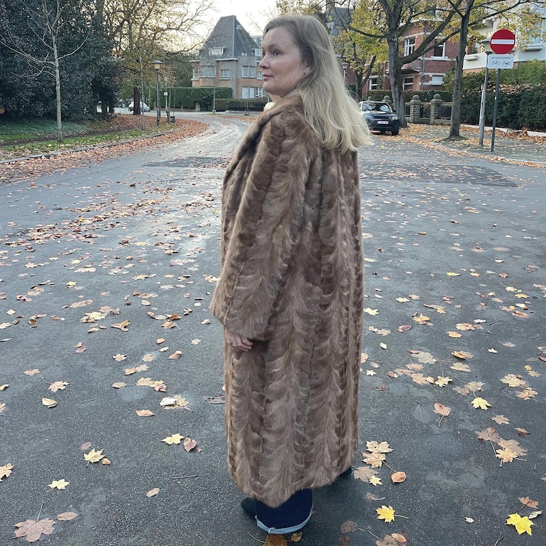 Pu&ograve; includere: Un lungo cappotto di pelliccia marrone con un motivo strutturato. Il cappotto arriva al ginocchio e ha un ampio colletto. La persona &egrave; in piedi su una strada con foglie cadute.