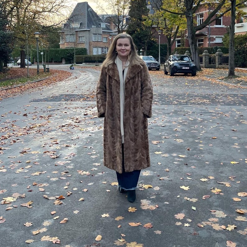 Pu&ograve; includere: Un lungo cappotto di pelliccia marrone con un motivo strutturato. Il cappotto &egrave; indossato sopra un top bianco e pantaloni blu scuro. La persona &egrave; in piedi su una strada coperta di foglie con edifici e auto sullo sfondo.