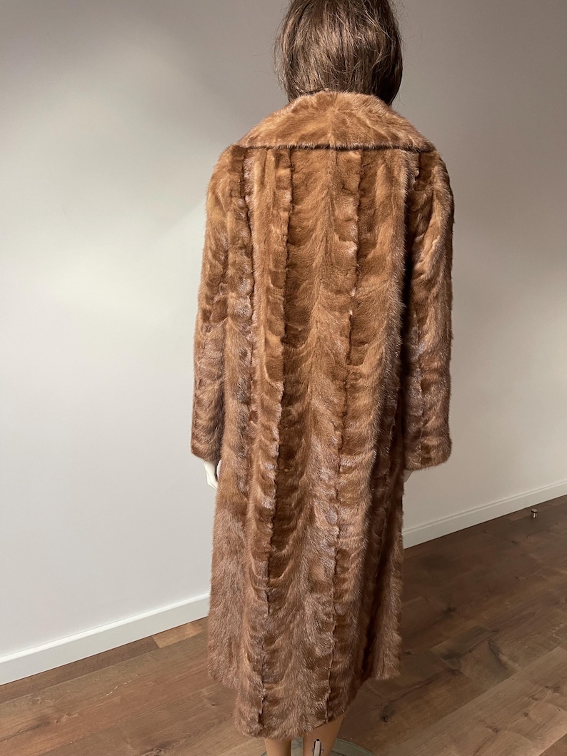 Pu&ograve; includere: Un cappotto di pelliccia marrone lungo fino a terra esposto su un manichino. Il cappotto ha un colletto a revers e maniche lunghe. La pelliccia &egrave; di un ricco e caldo colore marrone con un motivo strutturato. Il manichino &egrave; posizionato di spalle.