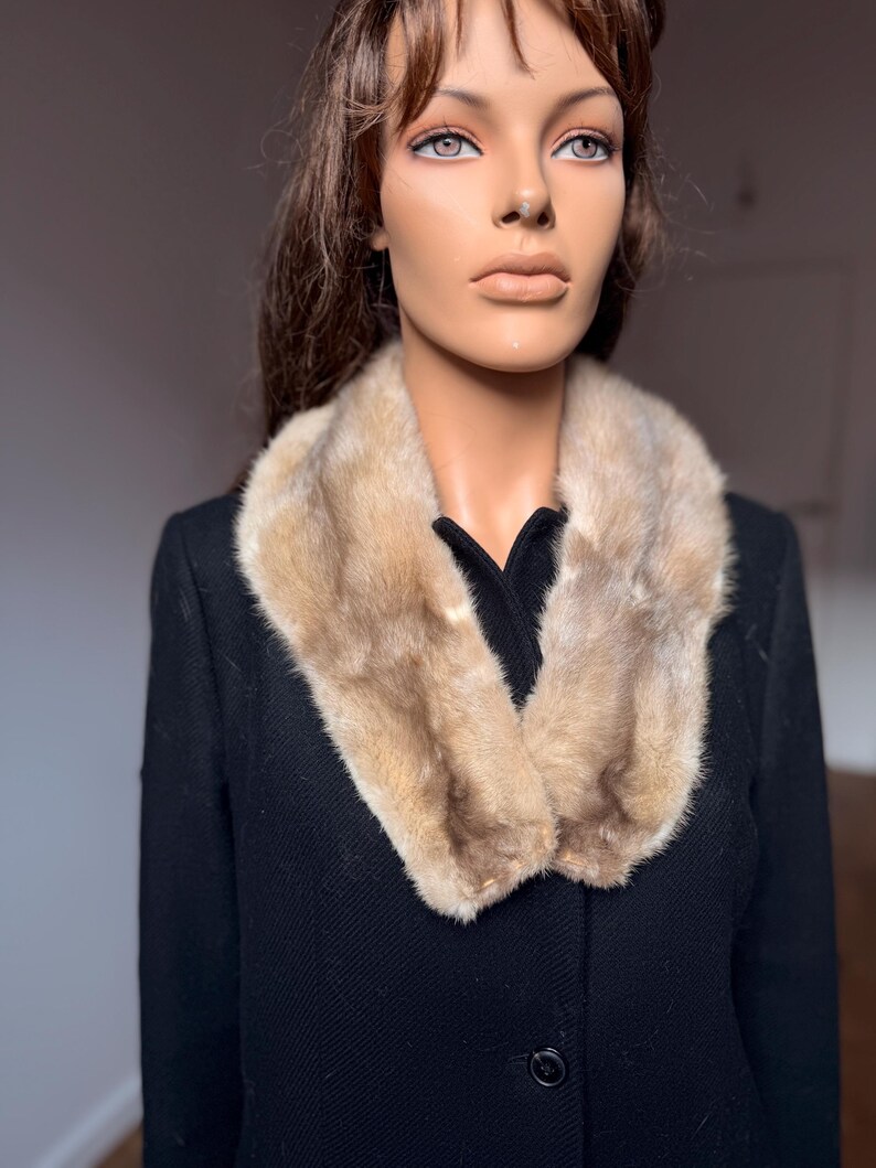 May include: A vintage black coat with a light brown fur collar. The fur collar is a light brown color and is draped around the neck of the coat. The coat has a classic design with a button closure.