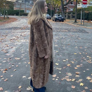 Pu&ograve; includere: Un lungo cappotto di pelliccia marrone con un ampio colletto. Il cappotto &egrave; indossato sopra jeans blu scuro e scarpe nere. La persona &egrave; in piedi su una strada coperta di foglie cadute.