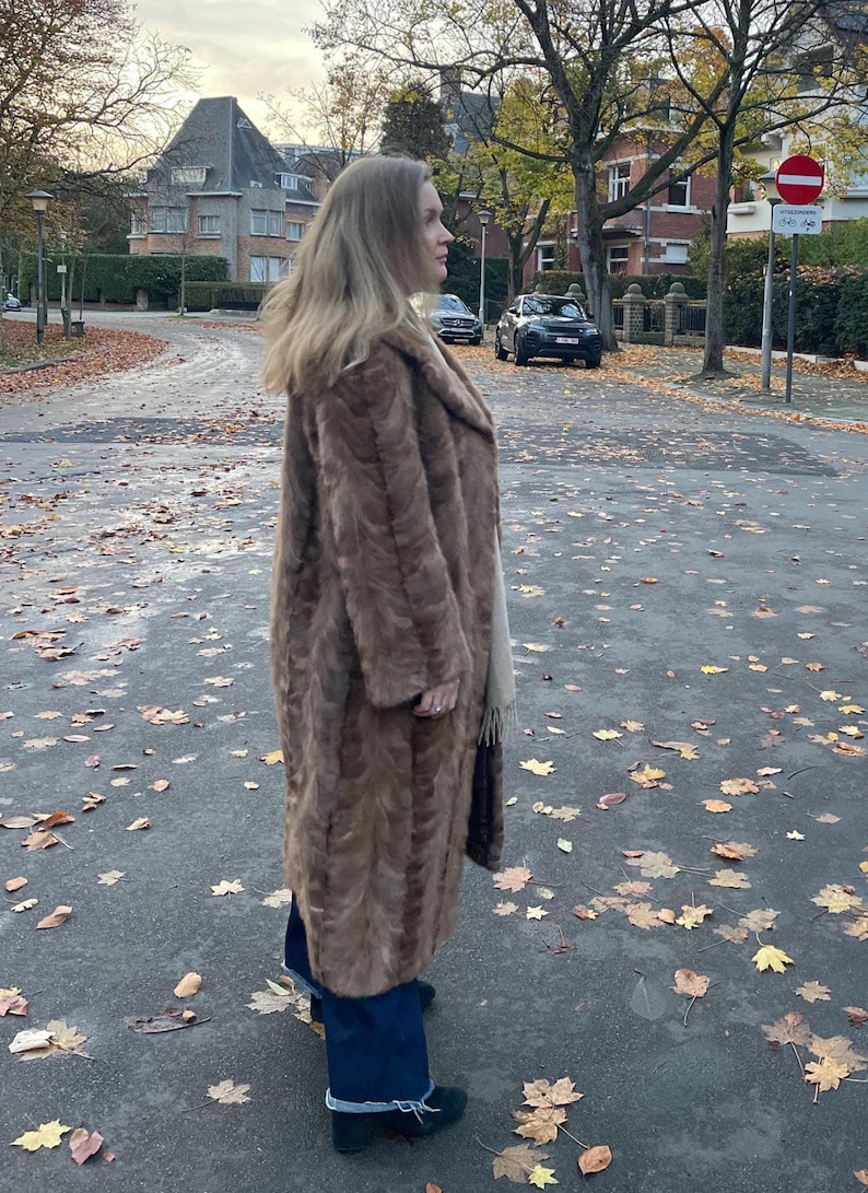 Pu&ograve; includere: Un lungo cappotto di pelliccia marrone con un ampio colletto. Il cappotto &egrave; indossato sopra jeans blu scuro e scarpe nere. La persona &egrave; in piedi su una strada coperta di foglie cadute.