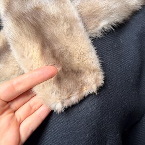 May include: Close-up of a coat with a light brown fur collar. The collar is soft and fluffy, contrasting with the dark navy blue fabric of the coat. The texture of the coat's fabric is visible.