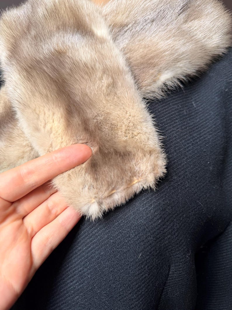 May include: Close-up of a coat with a light brown fur collar. The collar is soft and fluffy, contrasting with the dark navy blue fabric of the coat. The texture of the coat's fabric is visible.