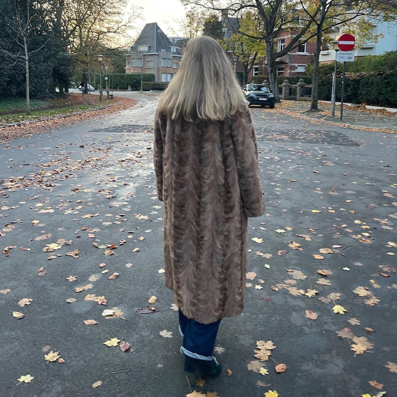 Pu&ograve; includere: Un lungo cappotto in finta pelliccia marrone con un motivo strutturato. Una persona cammina per una strada coperta di foglie cadute. La persona indossa pantaloni blu scuro e scarpe nere.