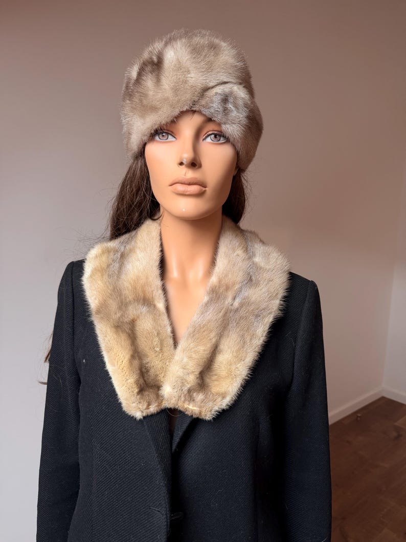 May include: A vintage fur hat and collar set. The hat is a light brown color with a rounded shape. The collar is a matching light brown fur, worn over a black coat. The mannequin is wearing the hat and collar.