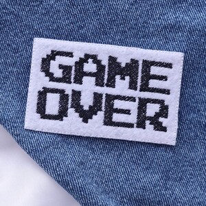 Puede incluir: Primer plano de una chaqueta vaquera con un parche blanco que dice "GAME OVER" en letras negras. La chaqueta es de color azul claro y tiene un forro blanco.