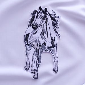 Può includere: Una toppa ricamata in bianco e nero raffigurante un cavallo in corsa. Il cavallo ha una criniera e una coda dettagliate, con un motivo maculato sul corpo. La toppa è su uno sfondo bianco.