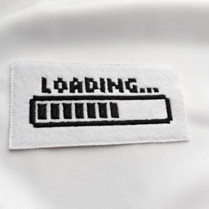 Peut inclure: Un écusson rectangulaire blanc avec le mot "LOADING..." brodé en noir. En dessous, une barre de progression noire, partiellement remplie. L'écusson est probablement destiné aux vêtements ou accessoires.