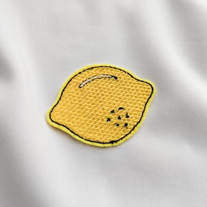 Może przedstawiać: Żółta, haftowana naszywka z cytryną z czarnym obrysem i małym białym zakrzywionym detalem. Naszywka ma teksturowany wygląd i jest umieszczona na białym tle.