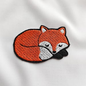 Op de afbeelding: Geborduurde patch met een slapende vos. De vos is oranje met witte accenten op het gezicht en de oren, en zwarte poten. De patch is zwart omlijnd en geplaatst tegen een witte achtergrond.