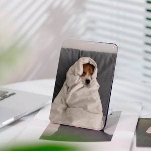 Könnte beinhalten: Ein Handy-Ständer mit einem Beagle, der in eine weiße Decke gehüllt ist. Der Hund liegt auf einem grauen Kissen. Der Ständer steht auf einer weißen Oberfläche mit einem Laptop und anderen Gegenständen im Hintergrund.