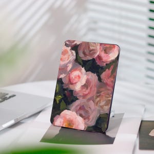 Könnte beinhalten: Ein rechteckiger Telefonständer mit einem Blumendesign aus rosa Rosen. Das Kunstwerk hat einen malerischen Stil mit sichtbaren Pinselstrichen und einem dunkelgrünen Hintergrund. Der Ständer steht auf einer weißen Oberfläche.