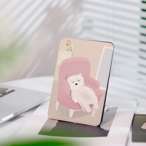 Könnte beinhalten: Ein rechteckiger Handyhalter mit einer Illustration eines weißen Hundes auf einem rosa Sessel. Der Hintergrund zeigt einen Raum mit einem Fenster und einer Einkaufstasche. Der Handyhalter liegt auf einer weißen Oberfläche.