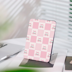 Peut inclure: Une housse de liseuse électronique rectangulaire présentant un motif répétitif de carrés à carreaux roses et blancs alternant avec des carrés à pois roses, chacun orné d'une petite illustration de deux fraises liées par un nœud rose.