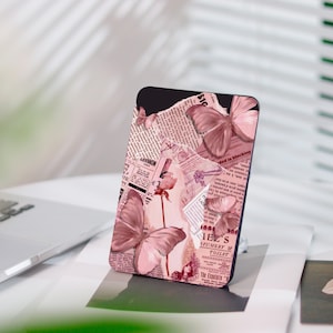 Può includere: Una custodia per telefono con un design di farfalle rosa e ritagli di giornale vintage. Il design include una rosa e vari elementi testuali, creando un'estetica romantica e artistica. La custodia è posta su uno sfondo neutro.