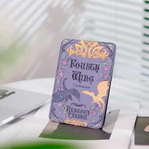 Könnte beinhalten: Ein lila Buchcover mit dem Titel "Fourth Wing" und der Autorin "Rebecca Yarros". Das Cover zeigt Illustrationen von Drachen, floralen Akzenten und keltischen Knotenmustern. Das Buch liegt auf einer weißen Oberfläche.