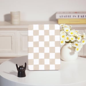 Könnte beinhalten: Ein Notizbuch mit beigem und weißem Schachbrettmuster steht auf einem weißen Tisch. Vor dem Notizbuch steht eine kleine schwarze Katzenfigur mit erhobenen Armen. Rechts befindet sich eine weiße Vase mit weißen und gelben Blumen.