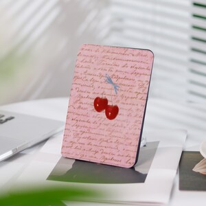 Könnte beinhalten: Eine rosa-weiße E-Reader-Hülle mit einem Vintage-Handschrift-Design und zwei roten Kirschen mit einem blauen Band.