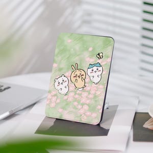 Könnte beinhalten: Eine grüne und rosa Handyhülle mit einem niedlichen Cartoon-Design von drei Tieren. Die Tiere sind ein weißes Kaninchen, ein gelbes Häschen und eine blaue Katze. Der Hintergrund ist ein grünes Feld mit rosa Blumen.