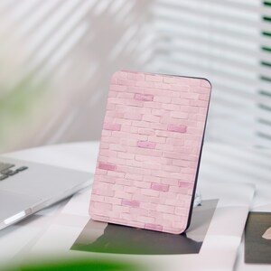 Könnte beinhalten: Eine rechteckige Handyhülle mit einem rosa Backsteinmuster. Die Hülle hat einen schwarzen Rand und liegt auf einer weißen Oberfläche. Das Backsteinmuster weist verschiedene Rosatöne auf, wodurch ein strukturiertes Aussehen entsteht. Die Hülle dient zum Schutz eines Telefons.