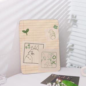 May include: A clear phone case featuring a notebook paper design with green leaf and flower stickers. The case has several cartoon Totoro illustrations in black outline. The word "OK" is also visible on one of the stickers.