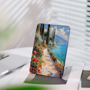 May include: A rectangular phone case displaying a landscape painting. The image shows a path with red roses and trees, leading to a blue lake and mountains under a sunny sky. The case is upright.