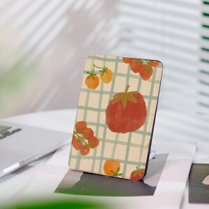 Può includere: Una custodia per telefono rettangolare con un design a pomodoro. La custodia ha uno sfondo color crema con un motivo a griglia verde e illustrazioni di pomodori rossi e arancioni. La custodia è esposta su una superficie bianca.