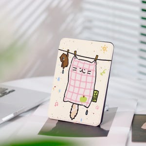Könnte beinhalten: Rechteckige Handyhülle mit einem Cartoon-Katzen-Design. Die Katze wird als Decke auf einer Wäscheleine mit einem kleinen Bären dargestellt. Die Hülle hat einen cremefarbenen Hintergrund mit rosa und grünen Akzenten.