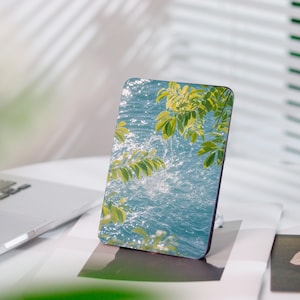 Könnte beinhalten: Eine rechteckige Handyhülle mit einem lebendigen Bild von Wasser mit Spritzern und grünen Blättern. Die Hülle hat eine glänzende Oberfläche und einen dunklen Rand. Das Design erinnert an Natur und Ruhe.