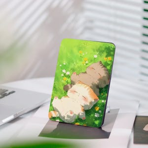 Könnte beinhalten: Eine rechteckige Handyhülle mit einer Illustration von zwei schlafenden Kätzchen in einem grasbewachsenen Feld mit kleinen gelben und weißen Blumen. Die Kätzchen sind hellbraun und orange, vor einem grünen Hintergrund.