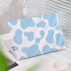 May include: A laptop with a light blue and white cow print case. The case covers the top of the laptop, with the Apple logo visible. The laptop is open, and the keyboard is visible. The background is white.