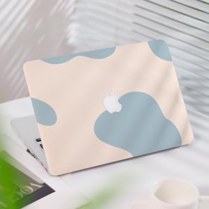 May include: A laptop with a light beige cover featuring soft blue abstract shapes. The Apple logo is visible. The laptop is on a white surface with a magazine and a cup in the background.