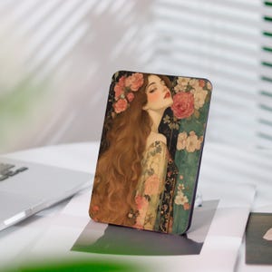 Könnte beinhalten: Ein rechteckiger Handyhalter mit einer romantischen Illustration einer Frau mit langen, rotbraunen Haaren und einem Blumenkranz. Das Kunstwerk zeigt Rosen und ein blumenmusterndes Kleid vor dunklem Hintergrund.