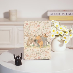 Könnte beinhalten: Rechteckige dekorative Holzplakette mit floralem Muster und einer Cartoon-Illustration von Winnie Puuh und seinen Freunden. Die Plakette steht auf einem weißen Tisch mit einer schwarzen Katzenfigur und einer Vase mit weißen Blumen.