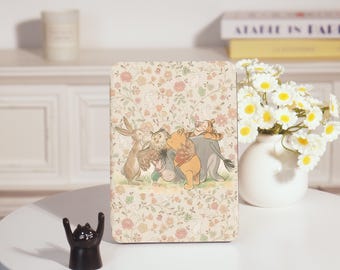 May include: A rectangular wooden plaque with a floral design and a cartoon illustration of Winnie the Pooh and friends. The plaque is on a white table with a black cat figurine and a vase of white flowers.