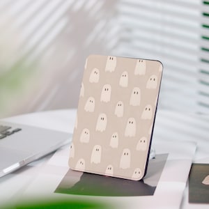 Può includere: Una custodia rettangolare con un motivo ripetuto di illustrazioni di fantasmi bianchi su sfondo beige. La custodia è in piedi su una superficie bianca, con un laptop visibile sullo sfondo. Il design è semplice e stravagante.