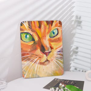 May include: A rectangular case featuring a vibrant, close-up painting of a cat's face. The artwork showcases a tabby cat with orange and yellow fur, bright green eyes, and long white whiskers. The case is set on a white surface.