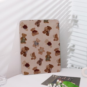 May include: A beige tablet case featuring a repeating pattern of teddy bears in various outfits. The bears are in shades of brown, wearing overalls, striped shirts, and sweaters. The case has a clear border.