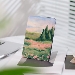 Può includere: Una custodia rettangolare per telefono con un dipinto di paesaggio raffigurante fiori rosa, colline verdi e alberi verde scuro sotto un cielo blu e rosa. La custodia è su una superficie bianca.