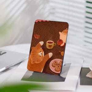 Könnte beinhalten: Eine braune und orange Katze mit weißer Brust und Pfoten liegt auf dem Rücken mit den Pfoten in der Luft. Die Katze liegt auf einer braunen Oberfläche mit einer Tasse Kaffee, einer Teekanne, einem Apfel und einem Teller Zimtschnecken. Der Hintergrund ist hellbraun.