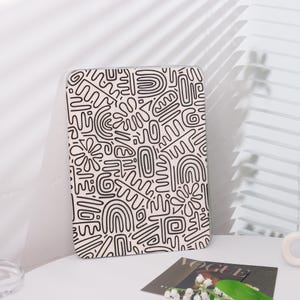 May include: A rectangular case with a cream background and a black abstract pattern. The design features various shapes and lines, including curved lines, ovals, and floral-like elements. The case is leaning against a white wall.
